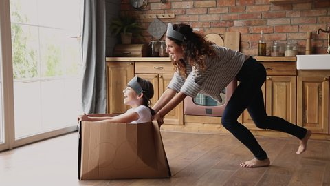 Active family play games at modern warm kitchen, young mom riding small daughter while she sitting inside of cardboard box, celebrate relocation day to new home, spend quarantine time creative concept