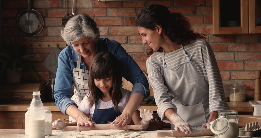 Three gen women in apron cooking together in kitchen, elderly granny, grown up daughter little granddaughter prepare pie, flattening dough enjoy process. Transfer skills from generation to gen concept Royalty-Free Stock Footage #1067355184
