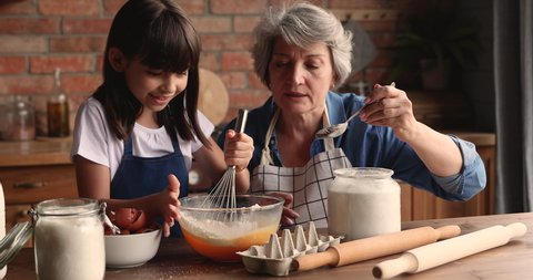 60s elderly female babysitter teach little girl to cook. Loving grandmother and small granddaughter prepare family recipe cake for event, stirring eggs make dough enjoy process. Hobby, cookery concept