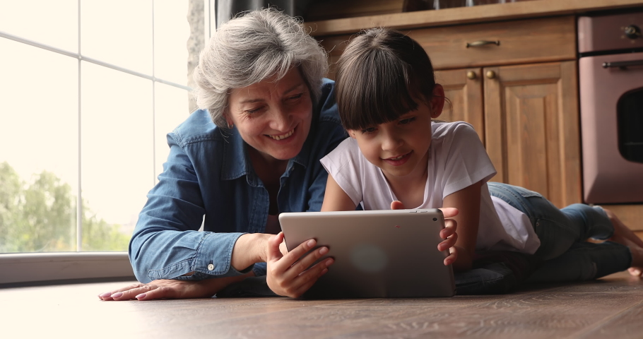 Elderly 50s grey-haired granny spend weekend with little cute grandkid family lying on floor using modern tech wireless tablet device, having fun online. Comfort and new cool mobile app usage concept Royalty-Free Stock Footage #1067355298