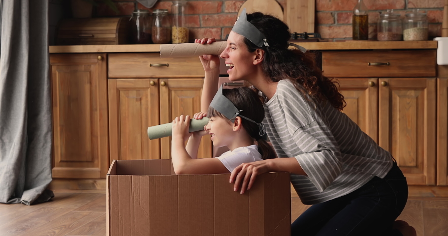 Small girl sit inside of carton box play pirates with mom in kitchen, wear drawn skull on caps, use paper tube like binocular watching into distance pretend be explorers. Playtime, fun at home concept Royalty-Free Stock Footage #1067355364