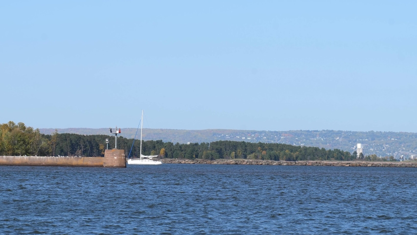 The ship channel at Wisconsin Point in Superior, with a sailboat at anchor near a concrete pier and Duluth, Minnesota past the trees in the distance, on a sunny day.