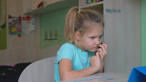 In the children's room with drawings on the wall, a girl with blond straight hair, a ponytail hairstyle, holds a finger in her mouth and looks into a smartphone, worries.