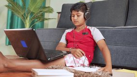 kid with headphone listening and noting down to book from laptop during virtual class from laptop at home - concept of online classroom, online education, technology and lifestyle