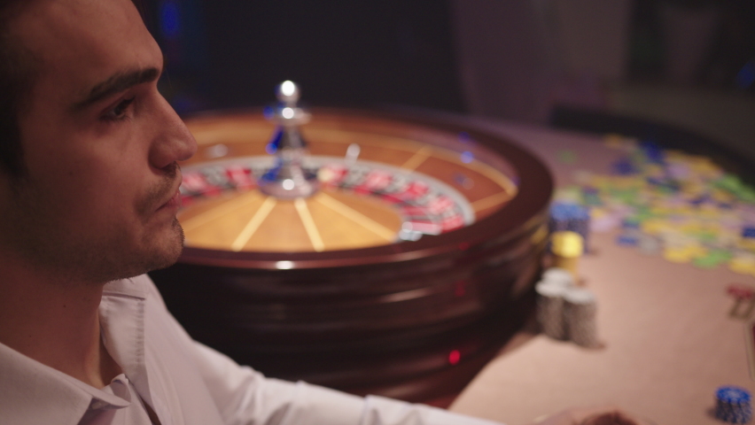 Nervous brunette man in white shirt drink whiskey and sitting alone behind the Roulette Table in the Casino while dealer takes away all chips from the table. Angry man after losing in casino. Royalty-Free Stock Footage #1067364443