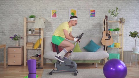 Funny man athlete from the 80's with a mustache falls off exercise bike on the couch