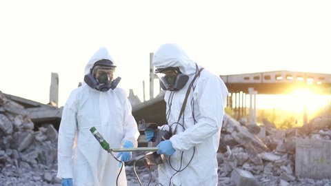 two scientists in protective suits and masks and a personal ionizing radiation dosimeter , walk measure radiation on the background of the ruins of a man-made disaster close up