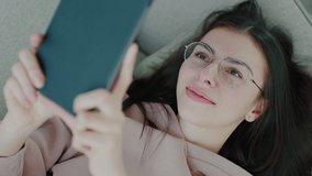 Close-up of a young Caucasian woman with glasses lying on a sofa, holding a tablet in her outstretched hand and having fun communicating via online video call.