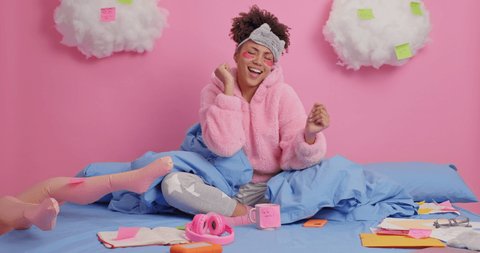 Curly beautiful African American woman moves and sings song has happy expression wears blindfold and nightwear poses on bed has working day at home surrounded by papers makes notes on stickers