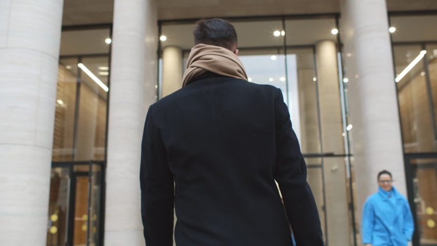 Back view of successful businessman entering business center. Confident male entrepreneur in coat and scarf walking through entrance doors in modern office building or apartment house