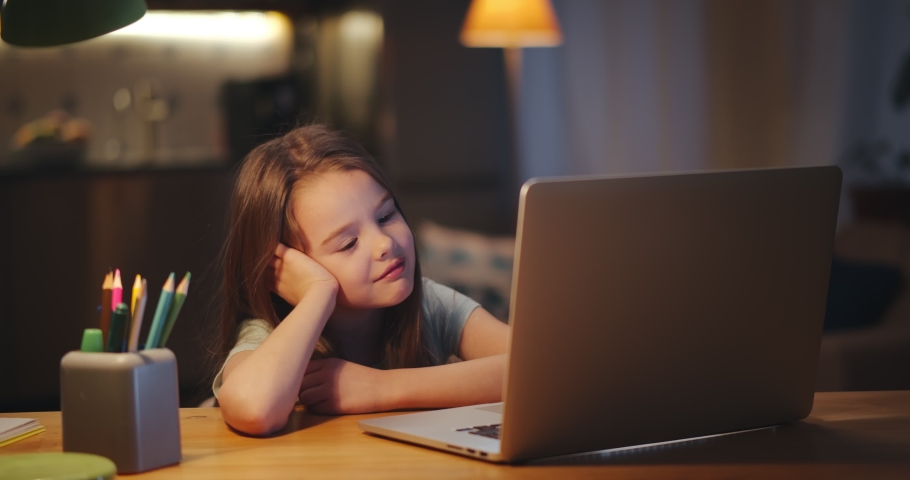 Preteen girl yawning doing homework at table with laptop in evening. Portrait of tired kid watching cartoons or studying online on computer sitting in dark living room | Shutterstock HD Video #1067390423