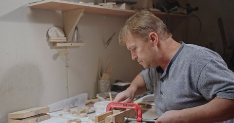 An adult carpenter in a tree workshop makes a wooden toy and smiles happily from the luck of trying to glue it together