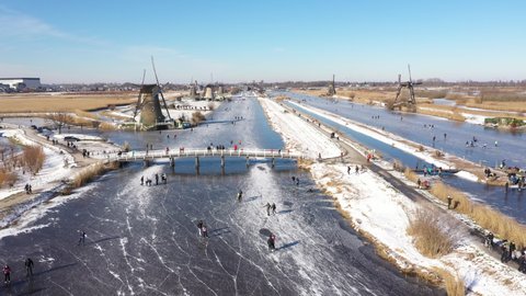 Drone flight over classic Winter landscape in the Netherlands. People ice skate on frozen canals past windmills, enjoying a traditional Dutch sport (a welcome break from Covid-19 lockdown)