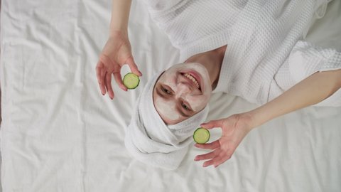 Body Love, Skin Care, After Bath, Cosmetics at Home, Caucasian Woman, Anti Aging. Caucasian woman in a bathrobe with a towel on her head lies on the bed and uses a cosmetic face mask and cucumber