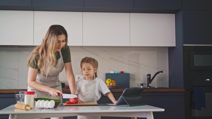 young mother in an apron in kitchen at table with ingredients, teaches young daughter with long hair to properly chop tomato, carefully, so as not to cut her fingers, with big knife