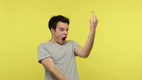 Rude impolite teenager in casual gray t-shirt showing middle finger fuck gesture at camera and angrily shouting, showing aggression, impolite behavior. Indoor studio shot isolated on yellow background