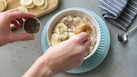 Adding chia seeds to oatmeal porridge bowl. Healthy breakfast food, diet meal of cooked oats, banana slices, peanut butter and seeds. Female cooking oats