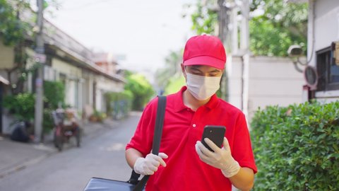 Asian delivery man in red uniform with face mask carrying bag of food delivering food to customer home during covid virus pandemic. He walking outdoor in village looking for customer home address.