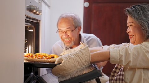 Asian old grandparents making pizza at home. Aged woman open the oven and bring the food out from machine. Elder man looking at meal and smell it with smile face, enjoy famiy activity together.