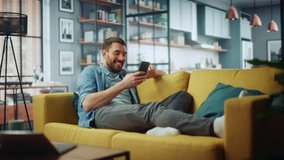 Happy Handsome Caucasian Man Using Smartphone in Cozy Living Room at Home. Man Resting on Comfortable Sofa. He's Browsing the Internet and Checking Videos on Social Networks and Having Fun.