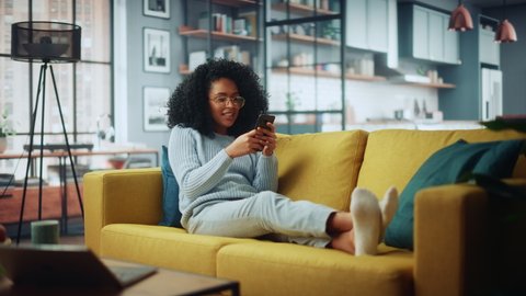 Happy Beautiful Latina Female Using Smartphone in Cozy Living Room at Home. Female Resting on Comfortable Sofa. She's Browsing the Internet and Checking Videos on Social Networks and Having Fun.