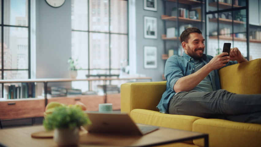 Happy Handsome Caucasian Man Using Smartphone in Cozy Living Room at Home. Man Resting on Comfortable Sofa. He's Browsing the Internet and Checking Videos on Social Networks and Having Fun. Royalty-Free Stock Footage #1067430695