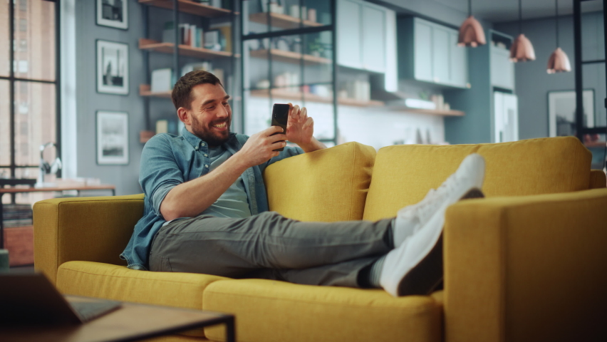 Happy Handsome Caucasian Man Using Smartphone in Cozy Living Room at Home. Man Resting on Comfortable Sofa. He's Browsing the Internet and Checking Videos on Social Networks and Having Fun.