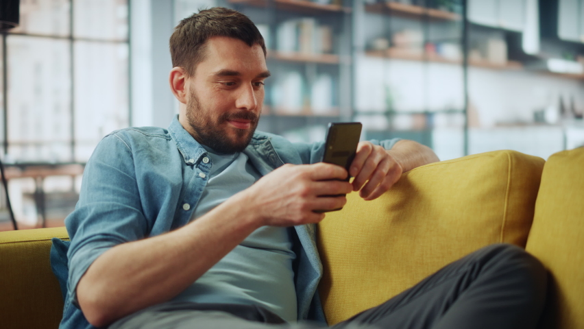 Happy Handsome Caucasian Man Using Smartphone in Cozy Living Room at Home. Man Resting on Comfortable Sofa. He's Browsing the Internet and Checking Videos on Social Networks and Having Fun. Royalty-Free Stock Footage #1067430701