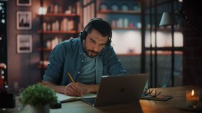 Handsome Caucasian Man With Headphones Talk on Video Conference Call on Laptop while Sitting in Dark Living Room in the Evening. Student Studying in Home School. Chat with Friends on Social Network.