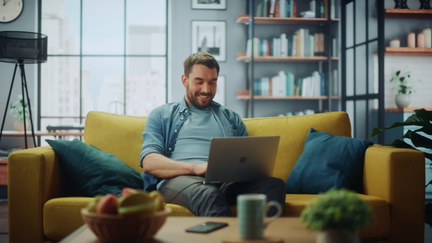 Handsome Caucasian Man Working on Laptop Computer while Sitting on a Sofa Couch in Stylish Cozy Living Room. Freelancer Working From Home. Browsing Internet, Using Social Networks, Having Fun.