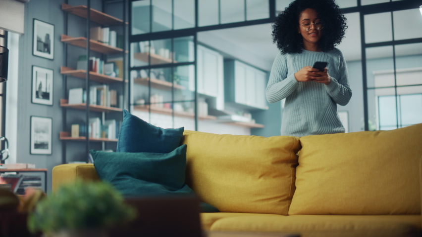 Happy Young Latina with Afro Hair Jumping Over on a Sofa Couch at Home while Using a Smartphone. Beautiful Diverse Multiethnic Hispanic Female Browsing the Web and Chatting with Friends. Royalty-Free Stock Footage #1067430989
