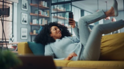 Happy Young Latina with Afro Hair Jumping Over on a Sofa Couch at Home while Using a Smartphone. Beautiful Diverse Multiethnic Hispanic Female Browsing the Web and Chatting with Friends.