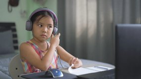 Asian child student or kid girl smile e-learning on computer notebook with wearing headphone and note on book for study online or people enjoy learn from home to play laptop for back to school on 4K