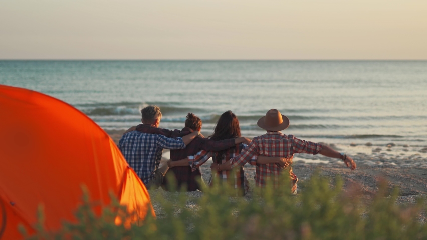rear view four best friends sitting at sunset on beach in front of sea and back to orange tent. people embracing and raising hands together during summer travel to seaside, enjoying beautiful sunset Royalty-Free Stock Footage #1067441546