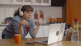 Young woman eating grapes in her home kitchen and watching video on the computer with headphones. Remote working concept. 
