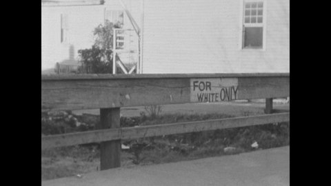 1930s: Woman walks down street past people standing near foliage. People hold branches. View of city street. View of church steeple. Railing with "For White Only" written on it.
