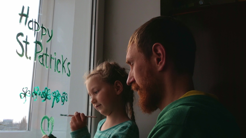 Drawing St. Patrick's Day Father with daughter painting green three-leaved shamrocks indoor, festive home decoration,family leisure. Drawing clover leaves on window glass. Stay home concept New normal Royalty-Free Stock Footage #1067449259