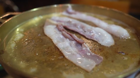 Close-up of Moo Ka Ta, Thai BBQ buffet with three pieces of sliced bacon are grilled on the BBQ grill pan with little boiling oil from the bacon in BBQ restaurant. Focus on the first piece of bacon.