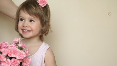 Mothers hands pin barrette with flower in girls hair. Child holds bouquet of pink carnations, looks at camera, smiles and smells flowers.