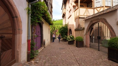 RIQUEWIHR, FRANCE - AUGUST 19, 2019: Walk at small alley of old village, through stone doorway, look to vineyards at hill side. Interesting corners of Riquewihr village, few unidentified people on way
