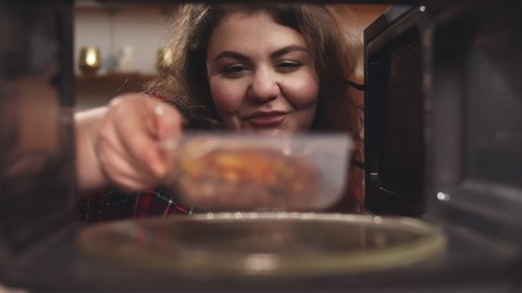 Beautiful young woman opening microwave oven and taking plastic container with heated meal. View from inside microwave of dinner warming and female taking it