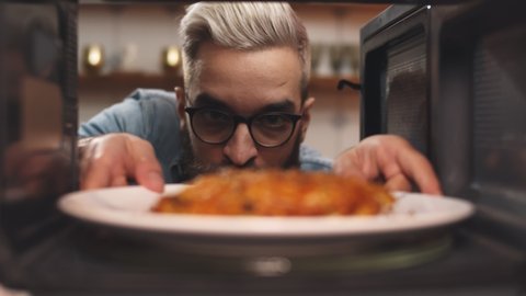 Man opening microwave oven door and taking warmed food. Young bearded guy warming cooked meal in plate in microwave for dinner. Kitchen appliance concept