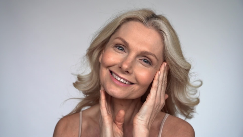 Happy 50s middle aged woman with flying blond hair touching facial skin looking at camera pampering in mirror. Old healthy skin care beauty concept, skincare treatment, anti age cosmetics. Slow motion | Shutterstock HD Video #1067463497