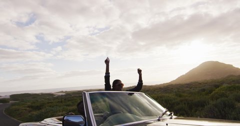 Diverse couple driving on sunny day in convertible car woman standing raising her arms in the air. summer road trip on a country highway by the coast.