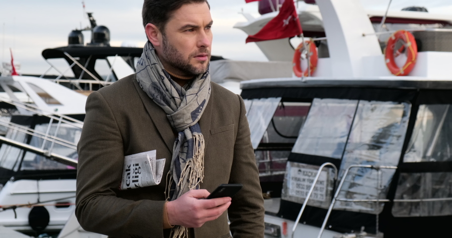 a man on the waterfront with a newspaper and a mobile phone is waiting for a business meeting with a business partner. businessman in an exclusive suit with a scarf near an  yacht,Turkey Istanbul Royalty-Free Stock Footage #1067467343