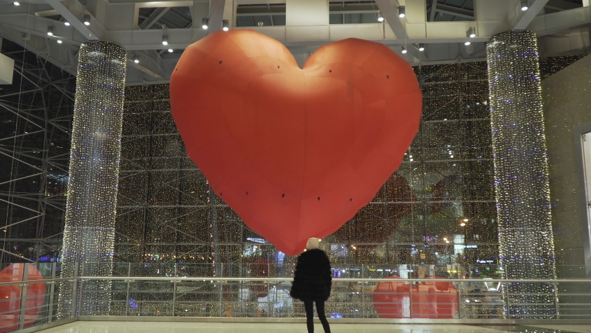 portrait of a young caucasian girl in a black fur coat and a pink hat in a shopping mall looks at a huge heart-shaped balloon on valentine