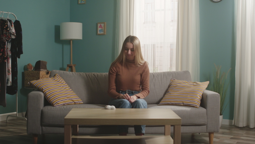 Young girl in brown sweater and jeans sits on beige sofa and plays with hamster running around coffee table. He catches him and kisses him.