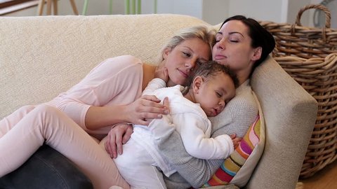 Same sex couple sleeping on their sofa with their baby son sleeping on one of their chests. 