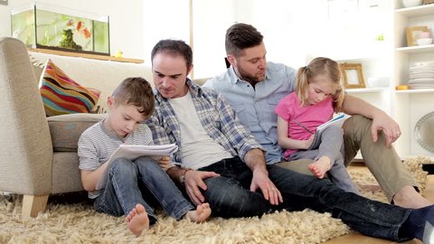 Male couple helping their son and daughter do their homework. They are sitting against a sofa in their home. Arkivvideo