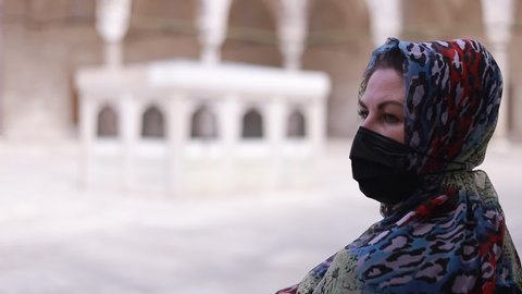 A woman in a shawl on the head and a protective mask walks in a mosque on a sunny summer day